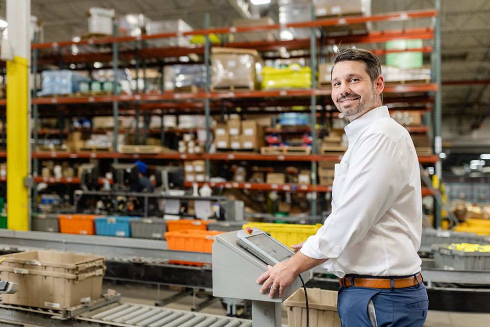 Industrial worker operating a control panel on a manufacturing floor with conveyor systems and warehouse inventory in the background