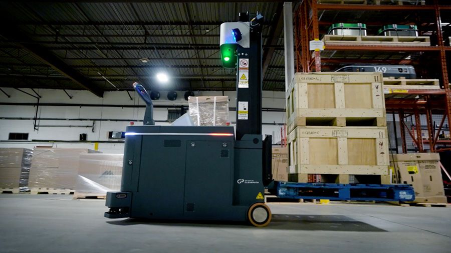 Autonomous mobile robot transporting a pallet of boxed materials through an industrial warehouse aisle with shelving and equipment