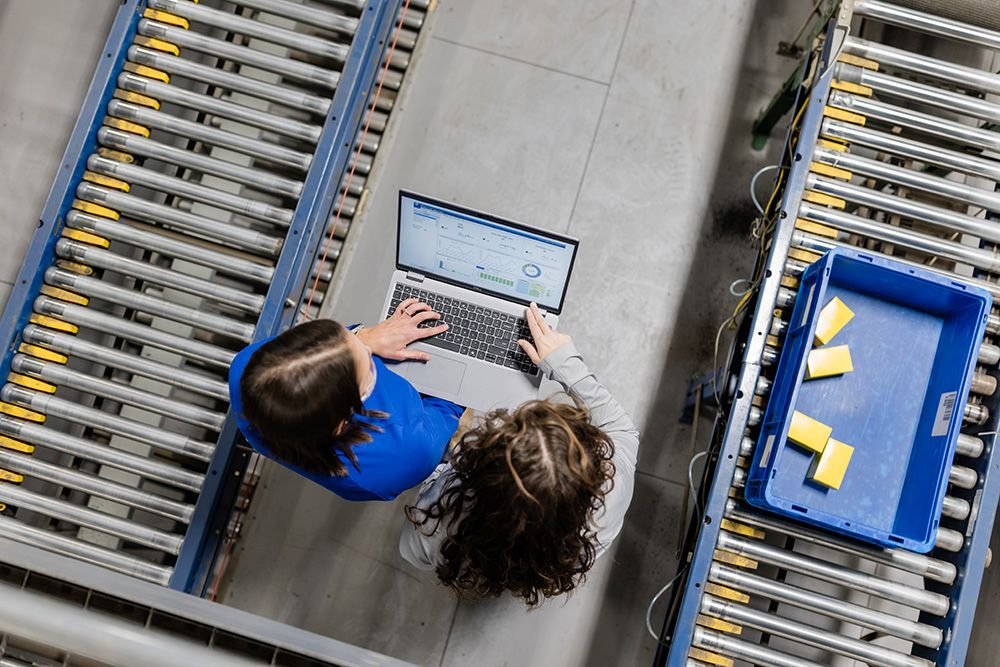 Overhead view of two people standing between conveyor lines in a warehouse while reviewing production data on a laptop next to a blue parts bin.